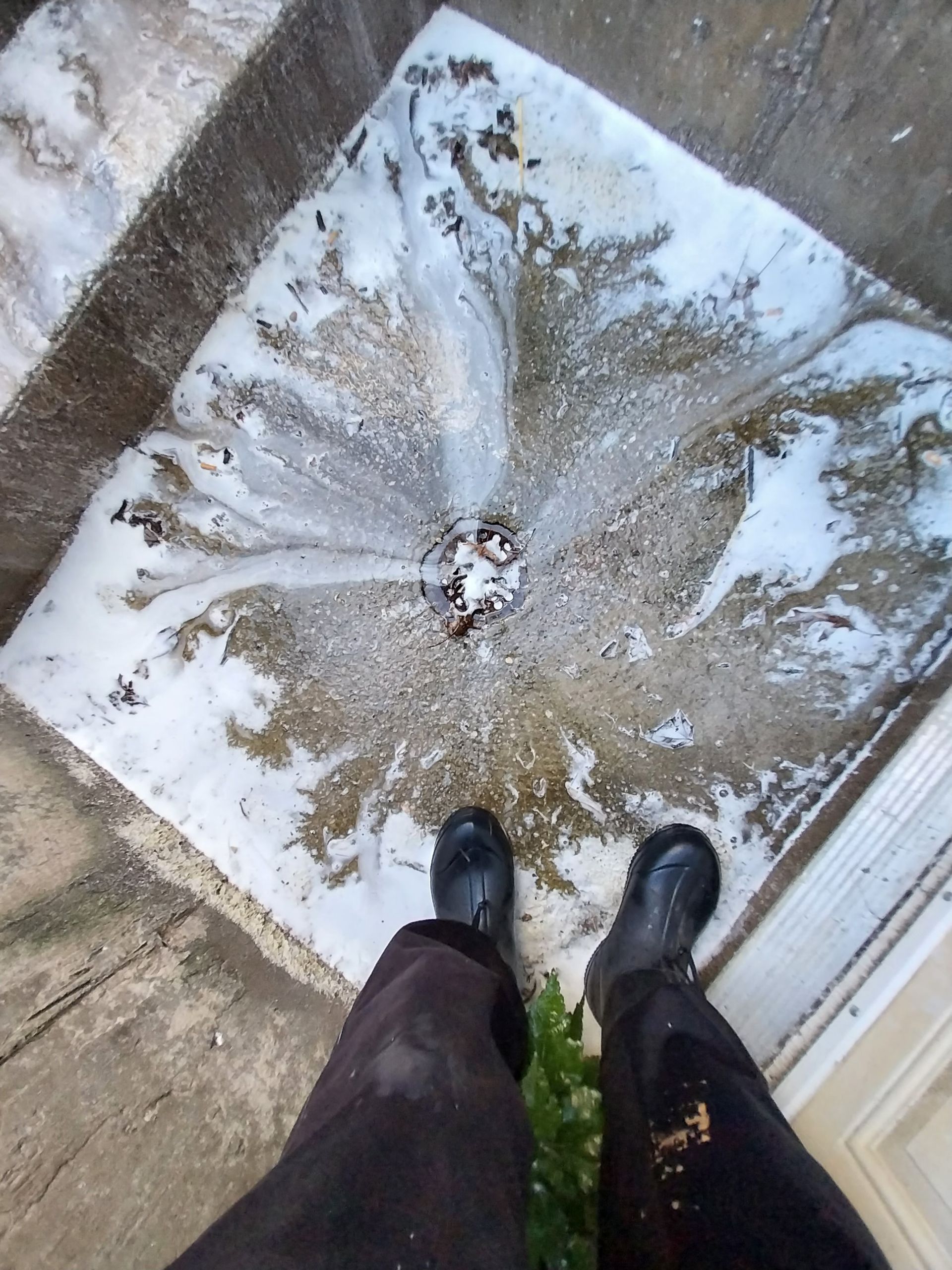 Person standing in front of a drain overflowing with foamy, white substance. They are wearing black boots and dark pants.