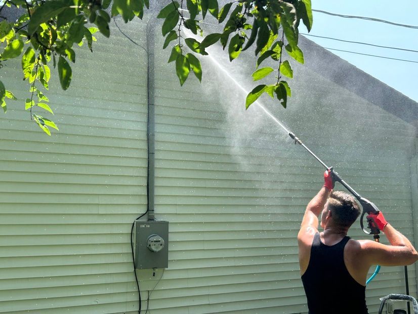 A person wearing a black tank top power washes a white-sided house, spraying water toward the wall under a leafy tree.