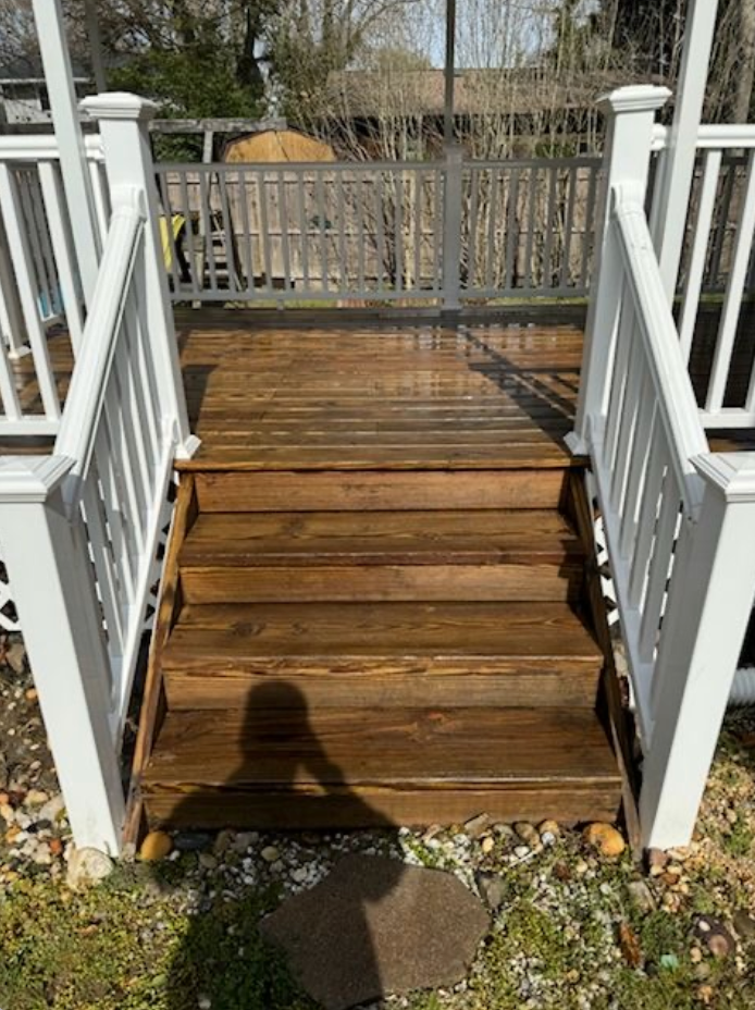 Wooden deck stairs with white railings. Sun casts a shadow across the stairs and grass.