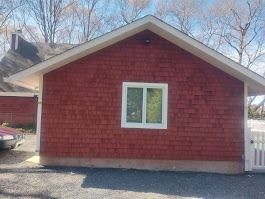 Red-shingled building with a white-framed window, set against a blue sky.  A white fence and partial car are visible nearby.