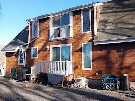 Brown shingled house with multiple windows and a white balcony. Outdoor area has assorted objects.