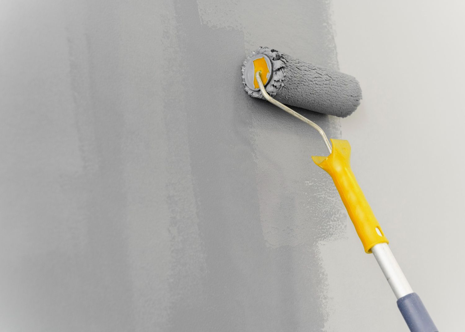 A paint roller, with a yellow handle, paints a gray wall.