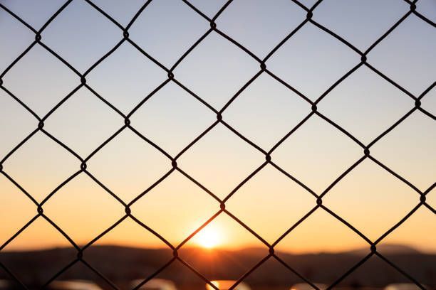 Chain link fence with sunset sky creating a silhouette effect behind the metal security mesh.