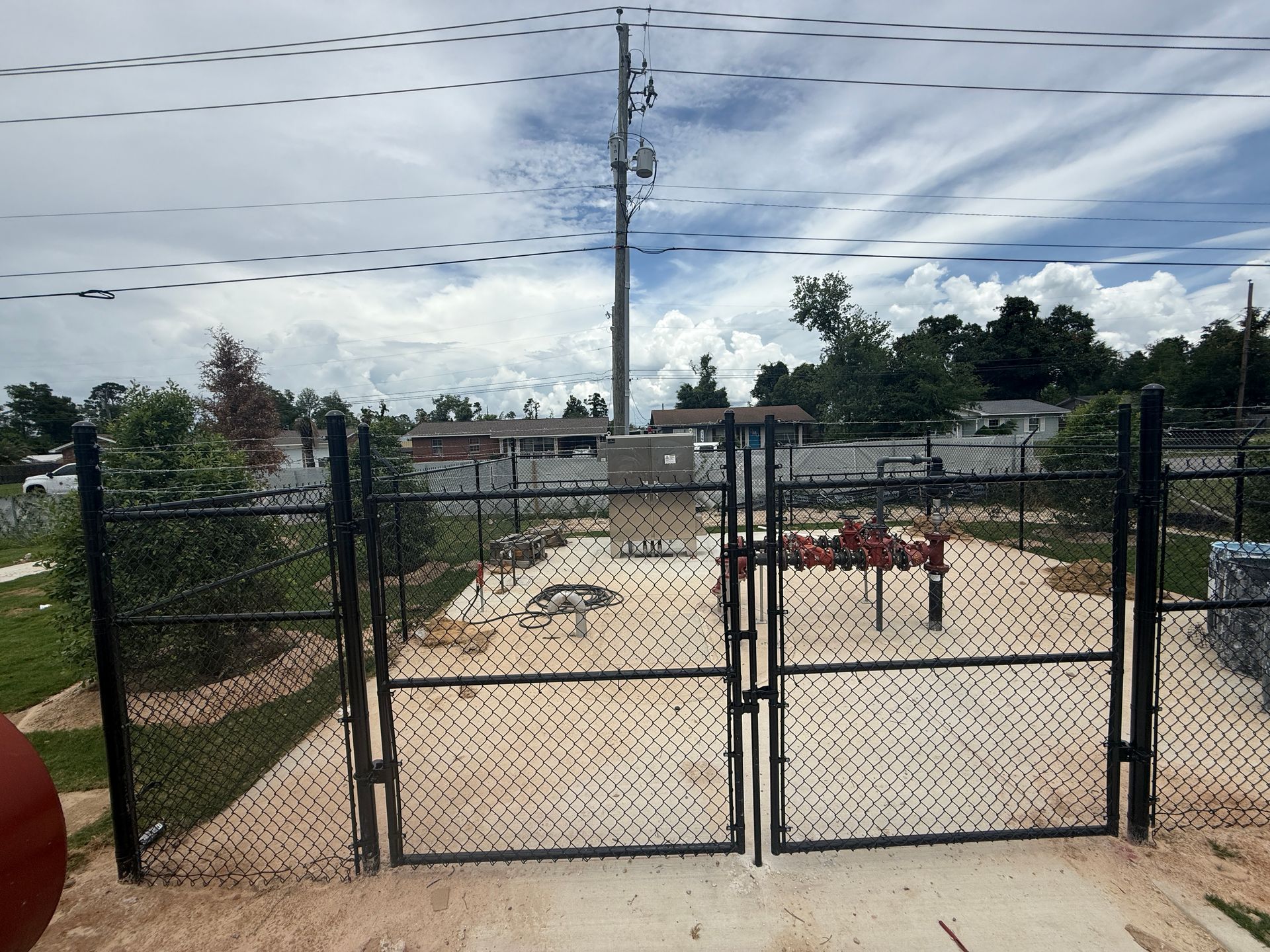 Fenced industrial site with black gate, concrete ground, equipment, power pole, and cloudy sky.
