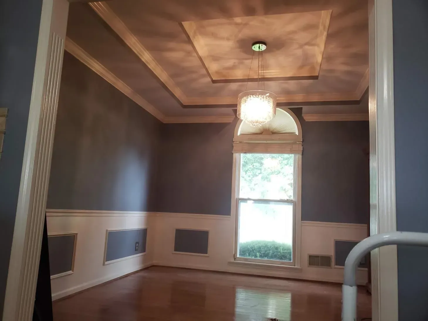 Empty dining room with blue walls, wainscoting, and a chandelier. Sunlight through a window, hardwood floors.