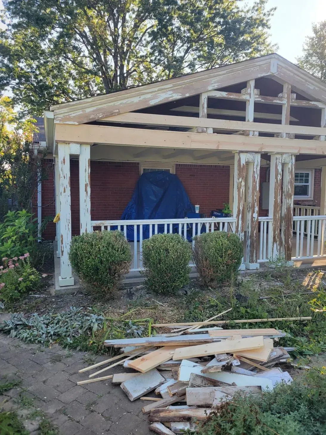 House porch under construction, peeling paint, wooden beams. Blue tarp covers something. Bushes in front.