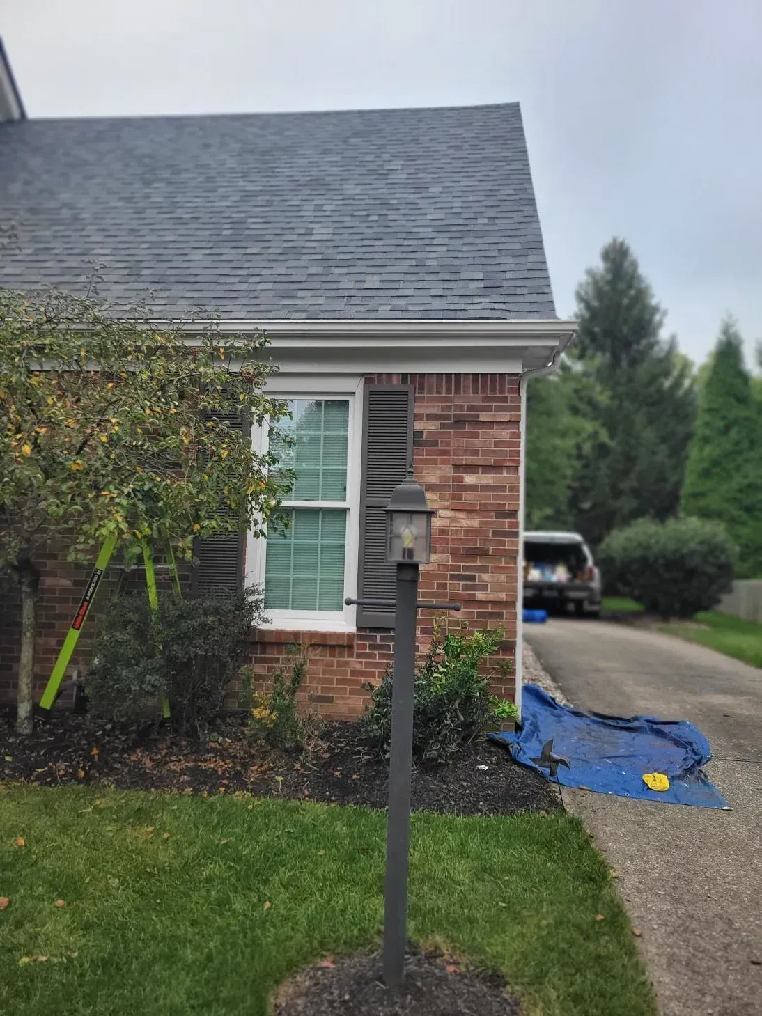 Red brick house corner with a window, black shutters, and a lamp post. A car is in the driveway.