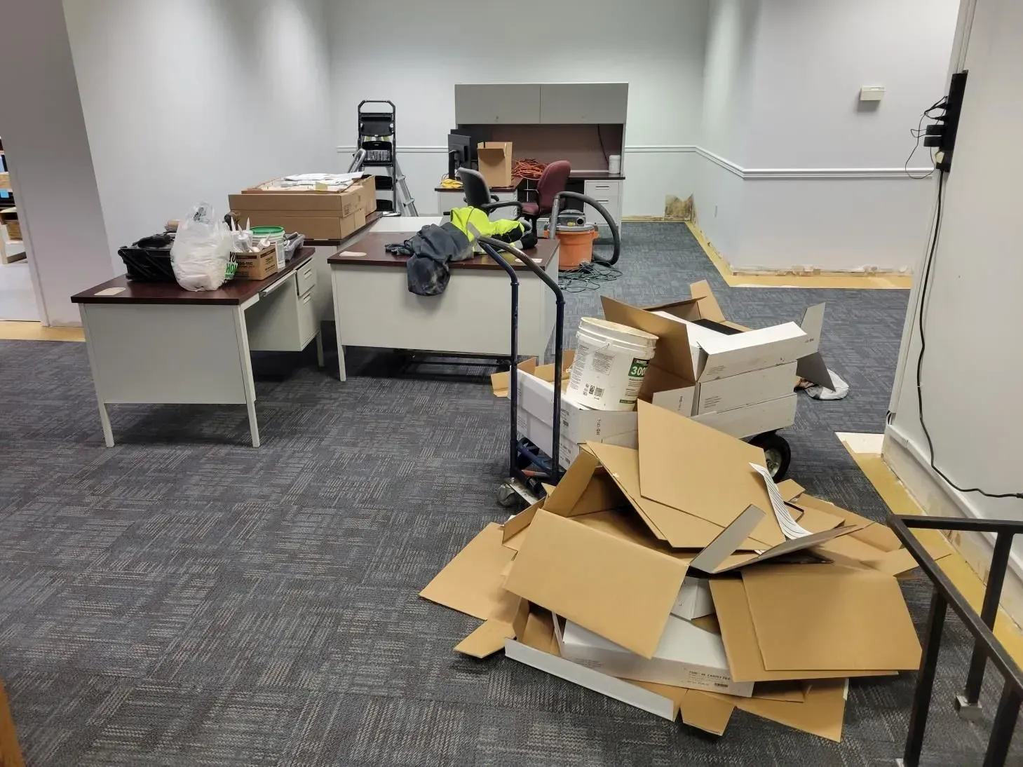 Office space with cardboard boxes, desks, and a vacuum, in disarray.