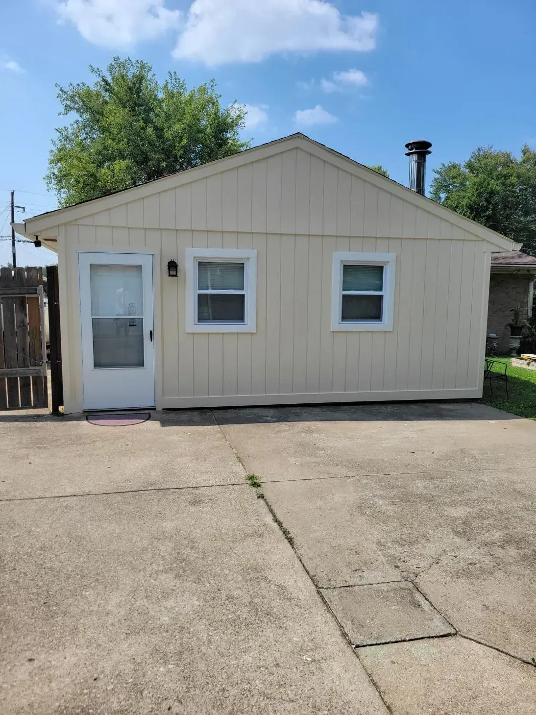 Small beige building with a door, two windows, and a chimney on a concrete driveway under a blue sky.