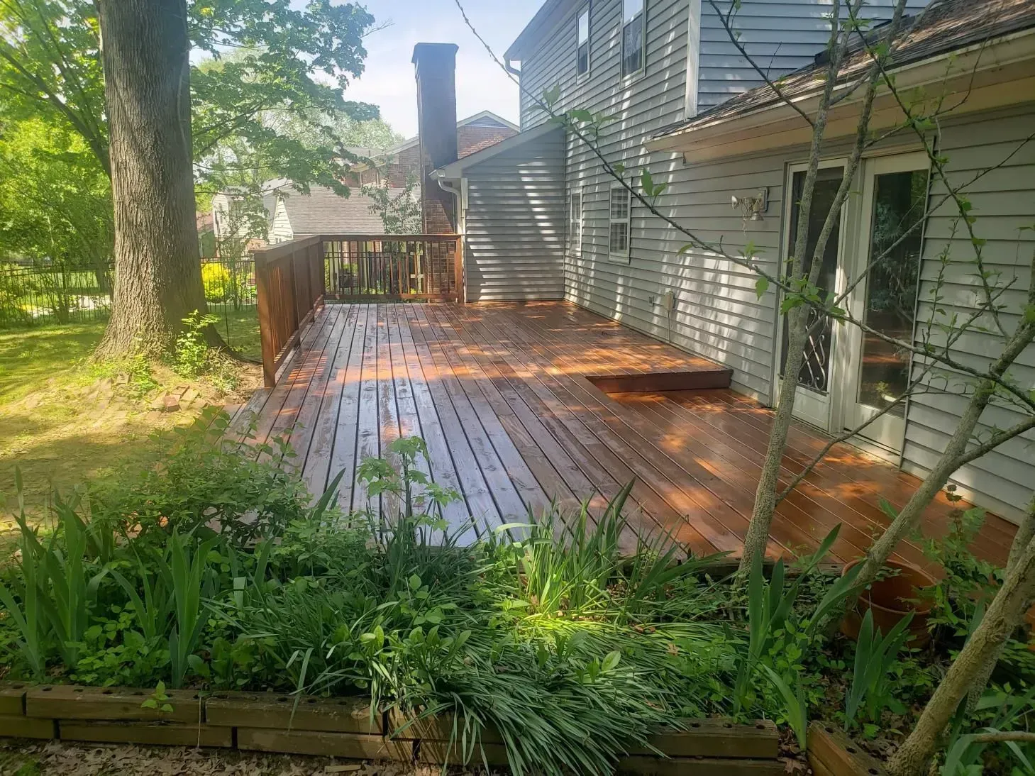 Wooden deck attached to a two-story house, surrounded by greenery.