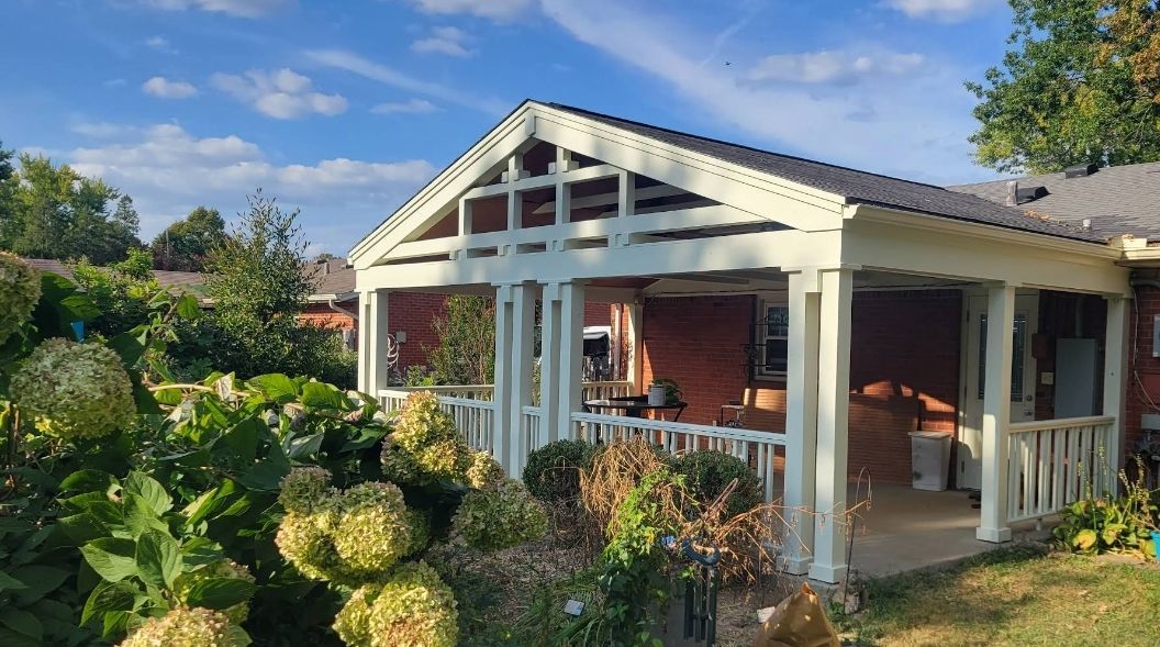 White porch with a black roof, next to a red brick house, and lush green garden.