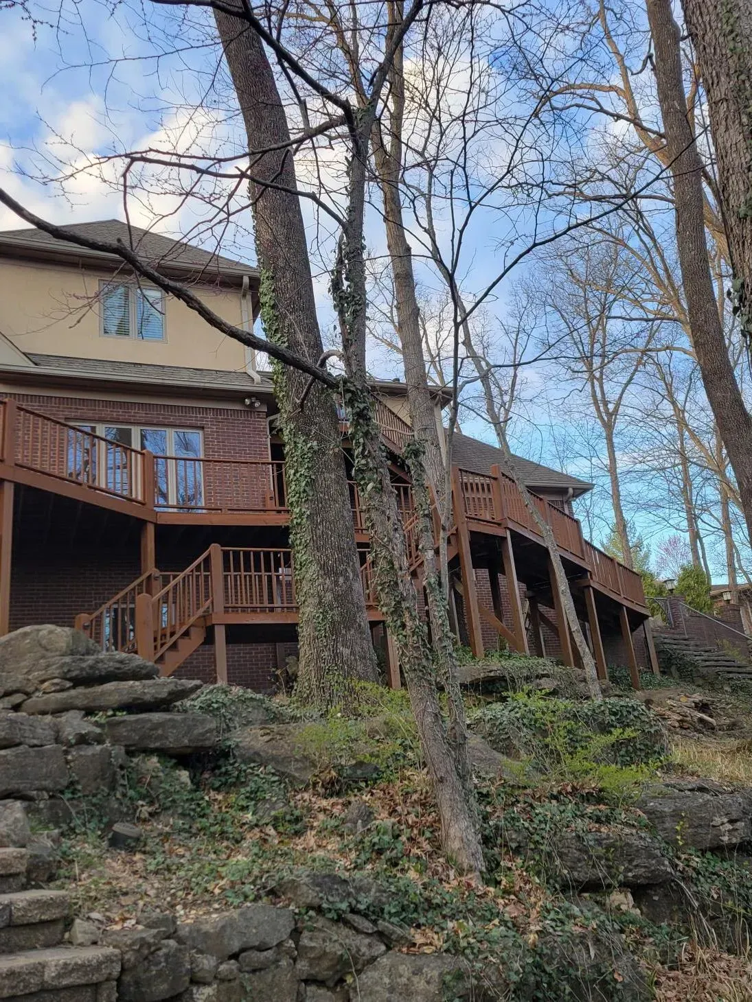 Two-story house with a wooden deck on a hillside, trees in front, and stone steps.