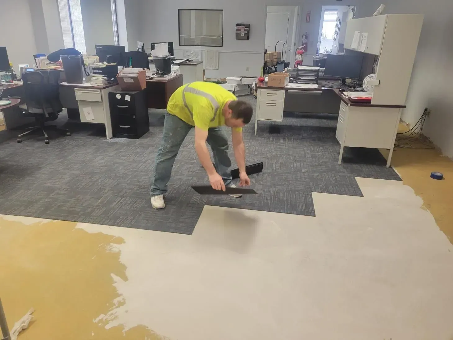 Person in a yellow vest installs gray carpet tiles in an office. The floor is partially covered.