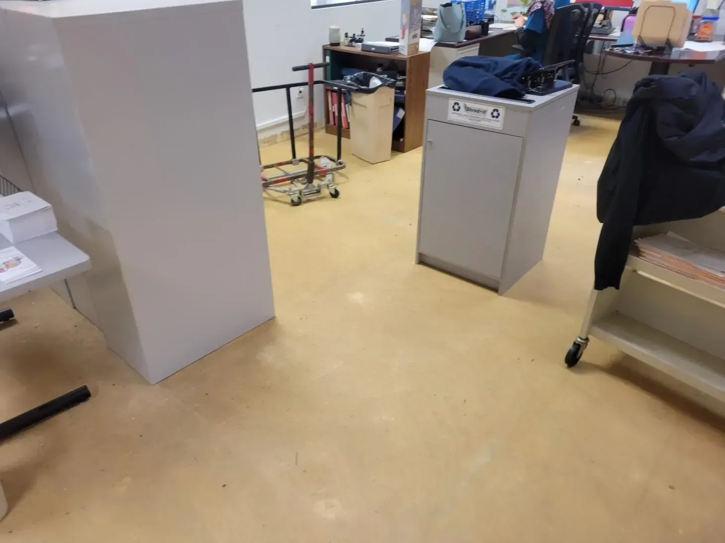Office interior with light-colored walls and floors. A jacket hangs on a cart. There are desks and storage.