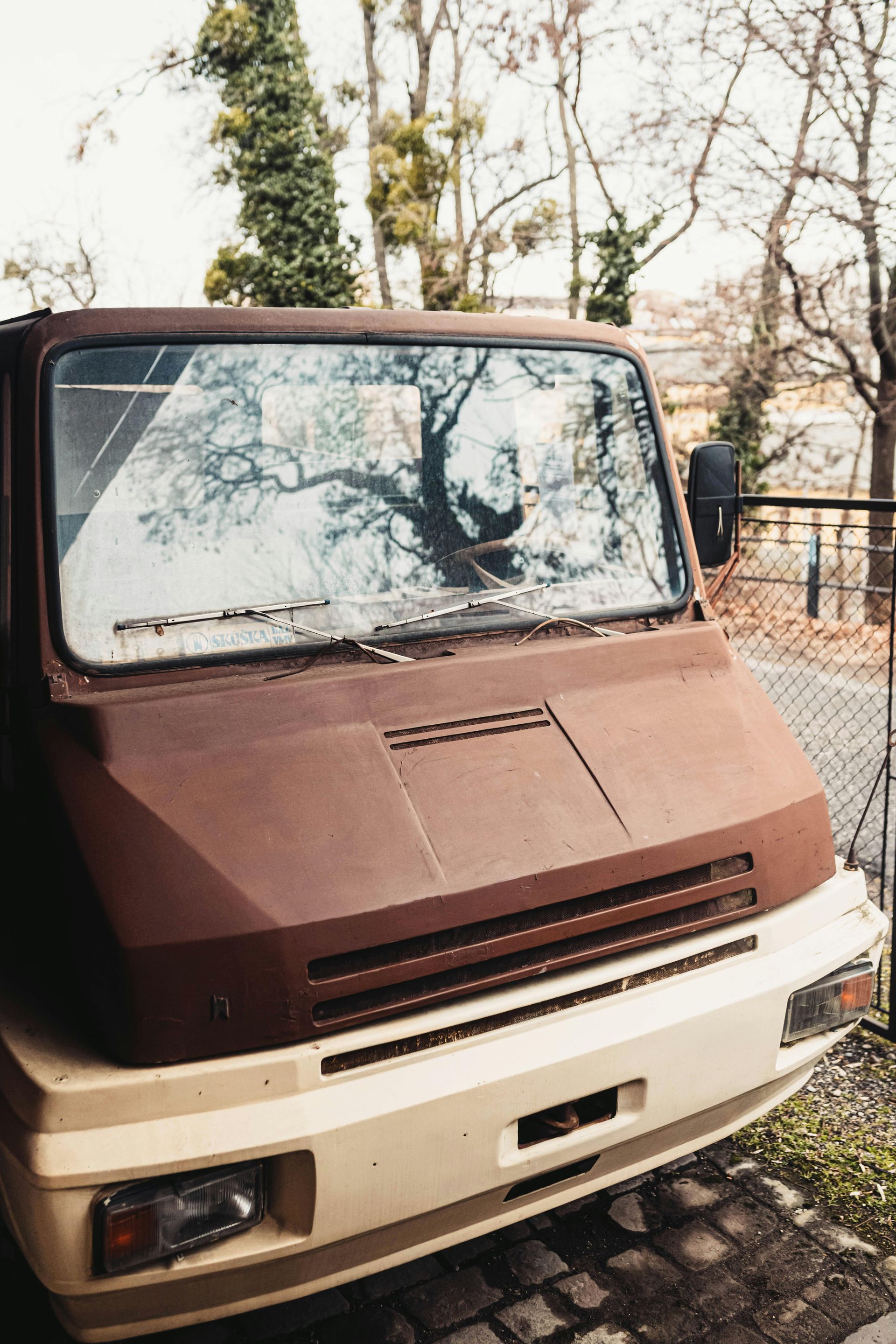 A weathered brown van parked under trees, showcasing a windshield with visible reflections and debris on the hood. 