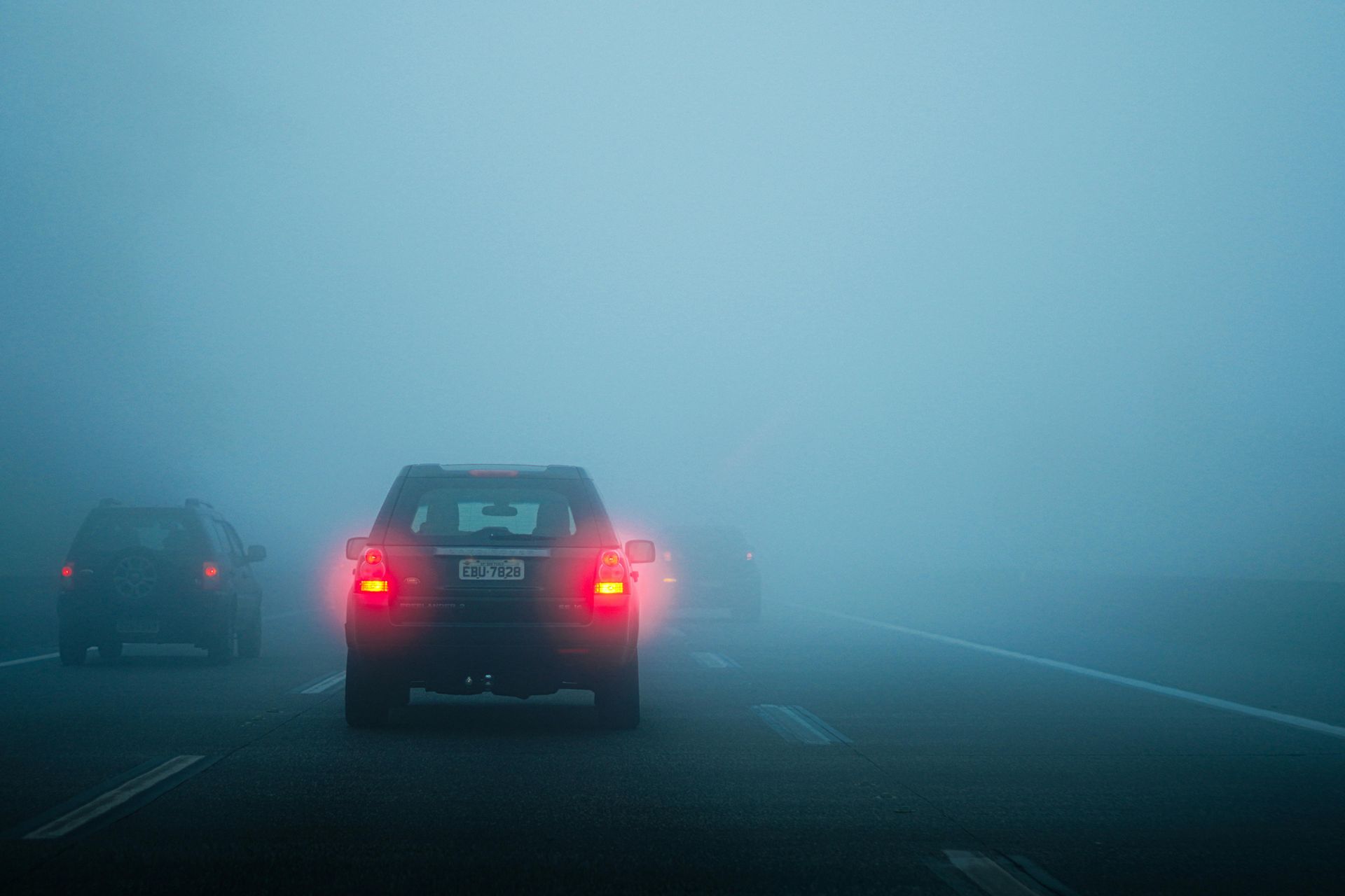 Cars driving on a highway in heavy fog, illuminated by red taillights.
