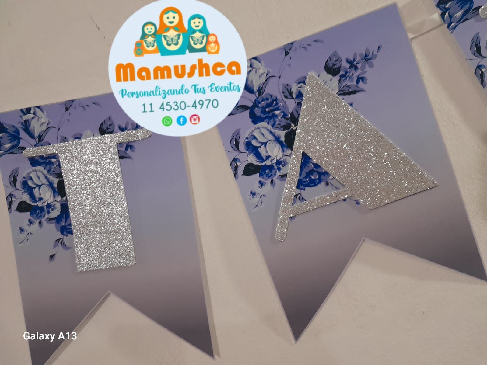 Pancarta con la palabra "TA" y letras brillantes sobre un fondo floral azul y banderines degradados. Logotipo de Mamushka en un círculo.