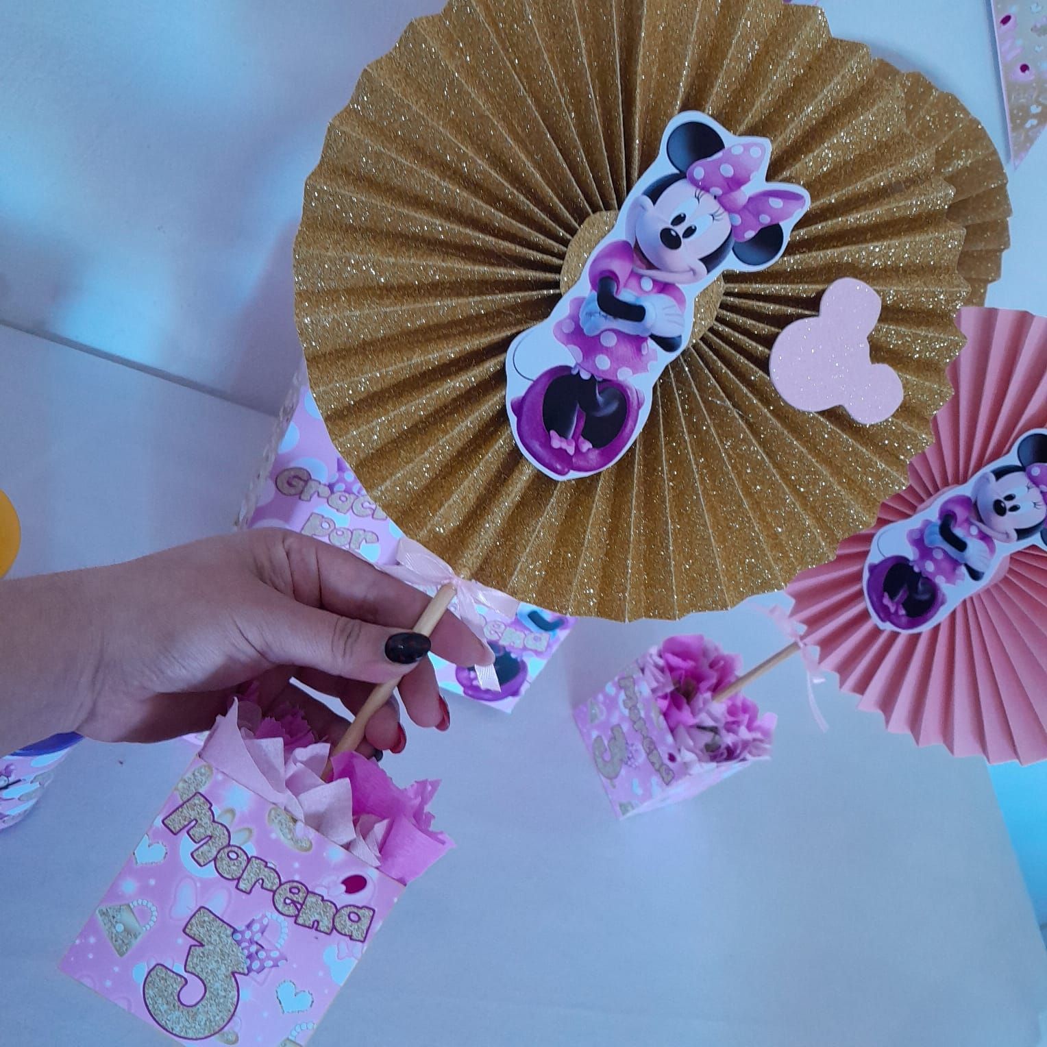 Persona sosteniendo una caja de regalo de fiesta de color rosa, probablemente para el tercer cumpleaños de un niño, con decoración de Minnie Mouse y abanicos de papel rosas y dorados.