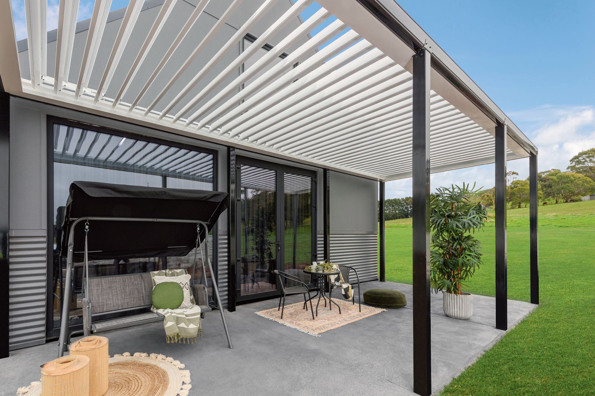 Patio with black pergola, white louvres, swing, small table, and greenery overlooking a lawn — Precision Patios Toowoomba In Toowoomba, QLD