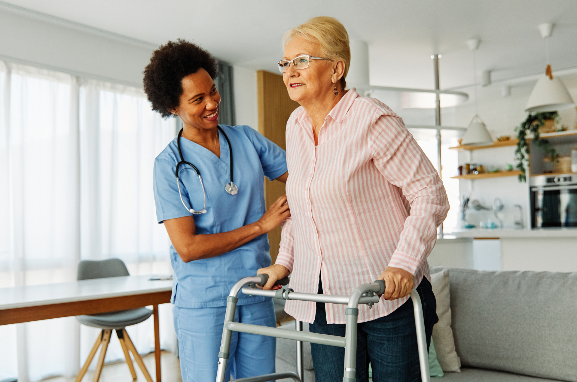 Nurse assists patient using a walker in a home. The patient wears glasses and pink shirt. The nurse smiles.