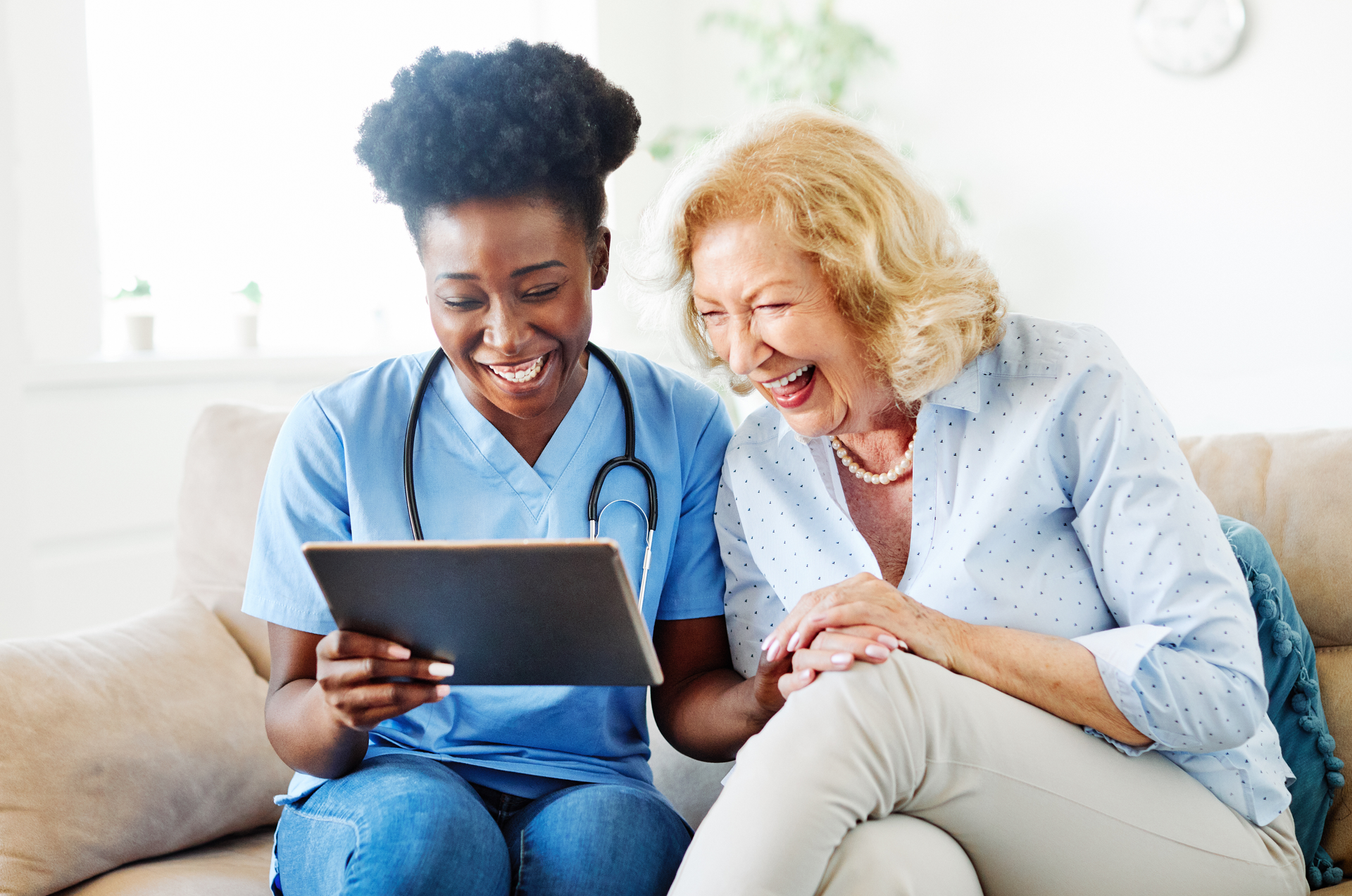 A healthcare provider and an older adult laugh while looking at a tablet together on a couch.