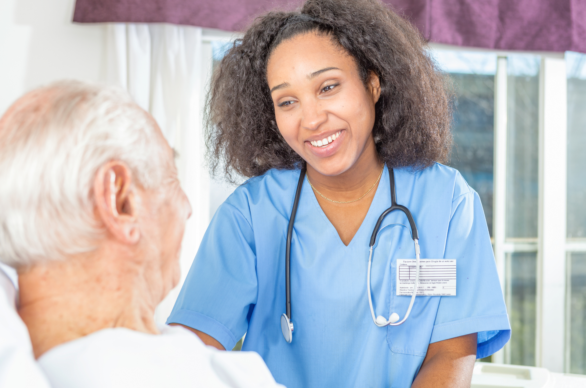 Nurse in blue scrubs smiles at elderly patient in a hospital room.