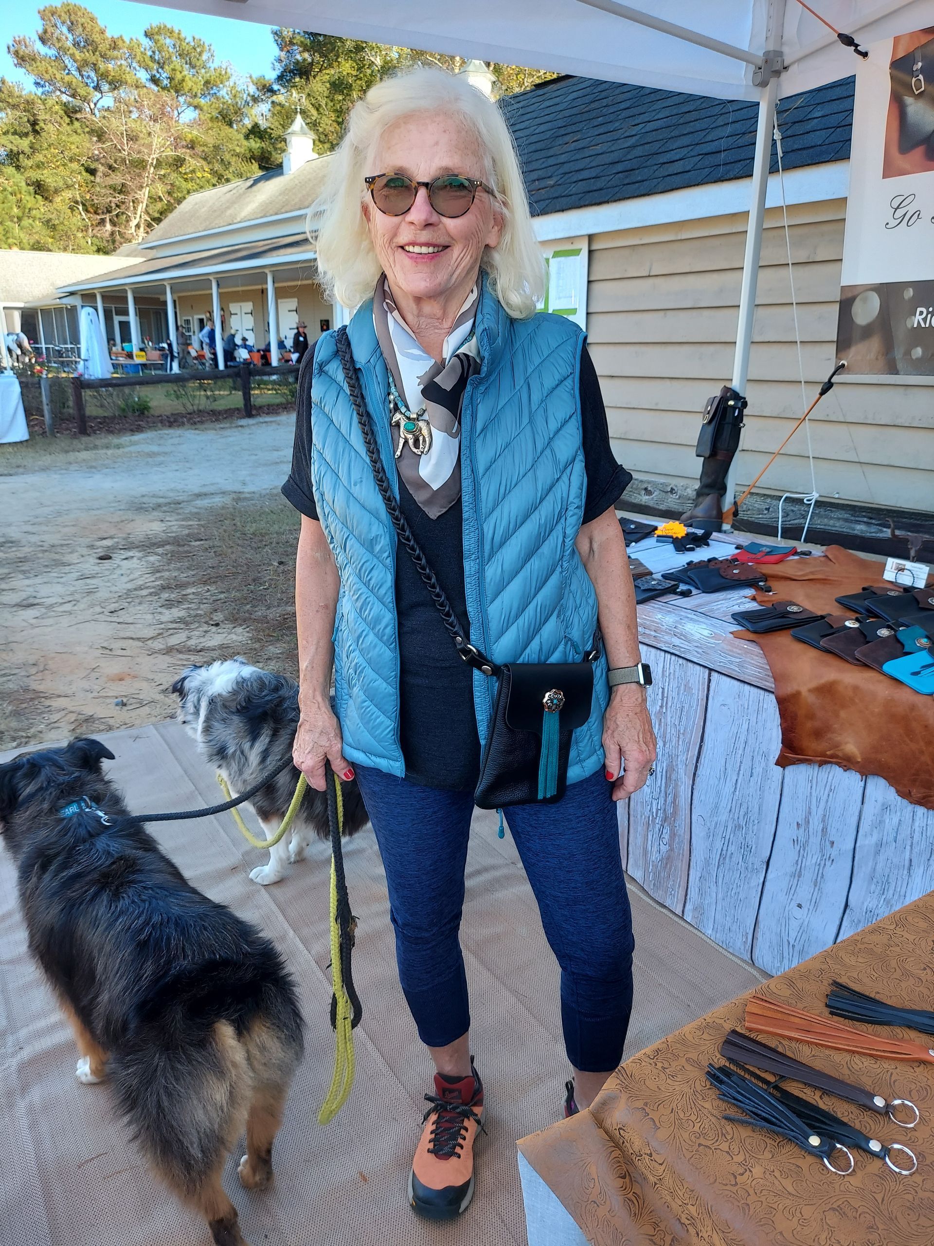 A smiling person wearing a blue quilted vest and sunglasses stands outdoors with two dogs on leashes near a market stall.