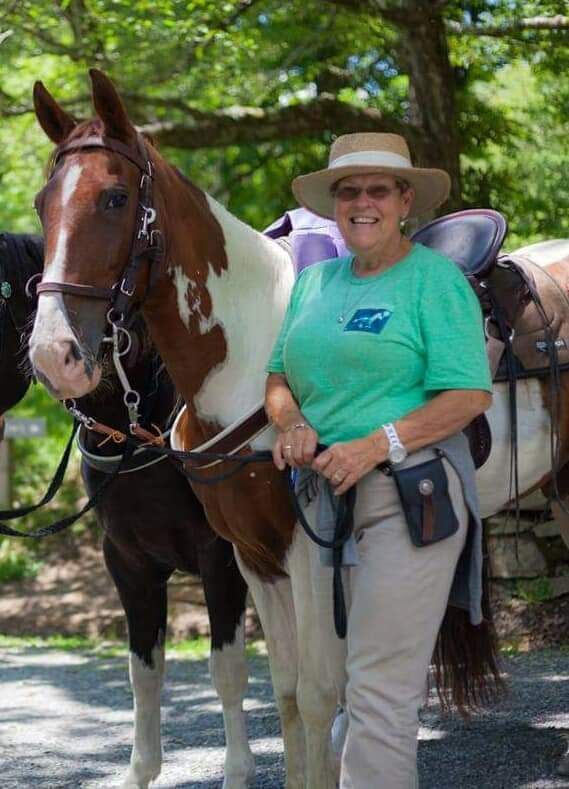 A smiling person in a green shirt and hat stands next to a saddled paint horse in a wooded outdoor setting.