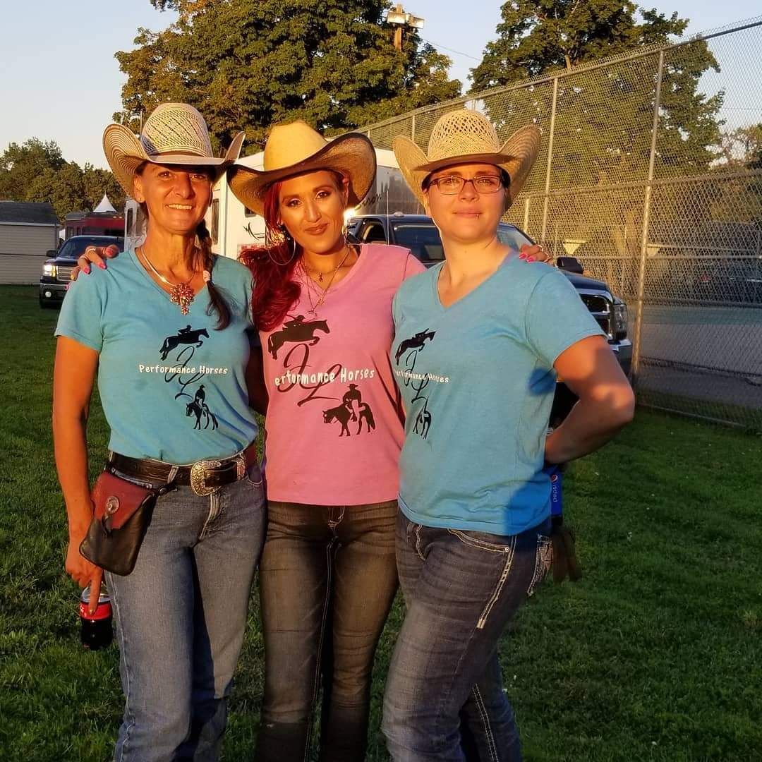 Three people wearing cowboy hats stand side-by-side in matching t-shirts and jeans outdoors on a grassy field.