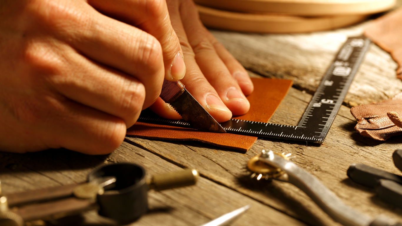 Person cutting leather with a knife and ruler on a wooden work surface.