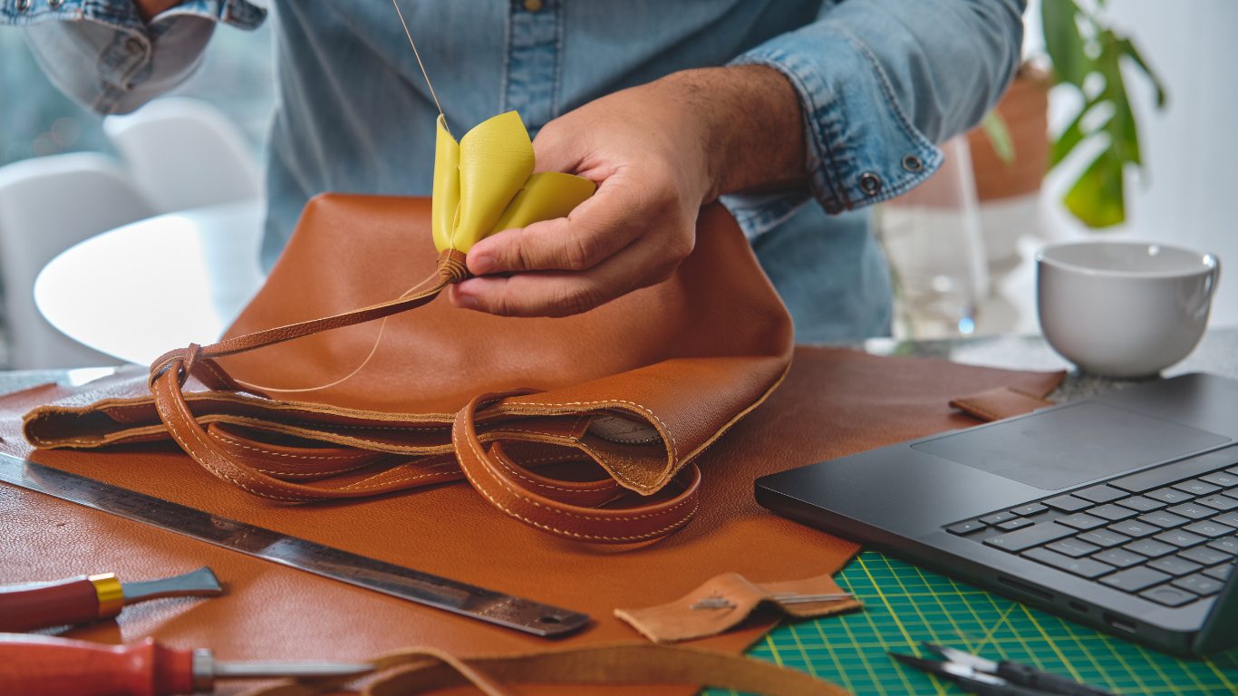 Person stitching leather bag, yellow flower detail, workspace with tools and laptop.