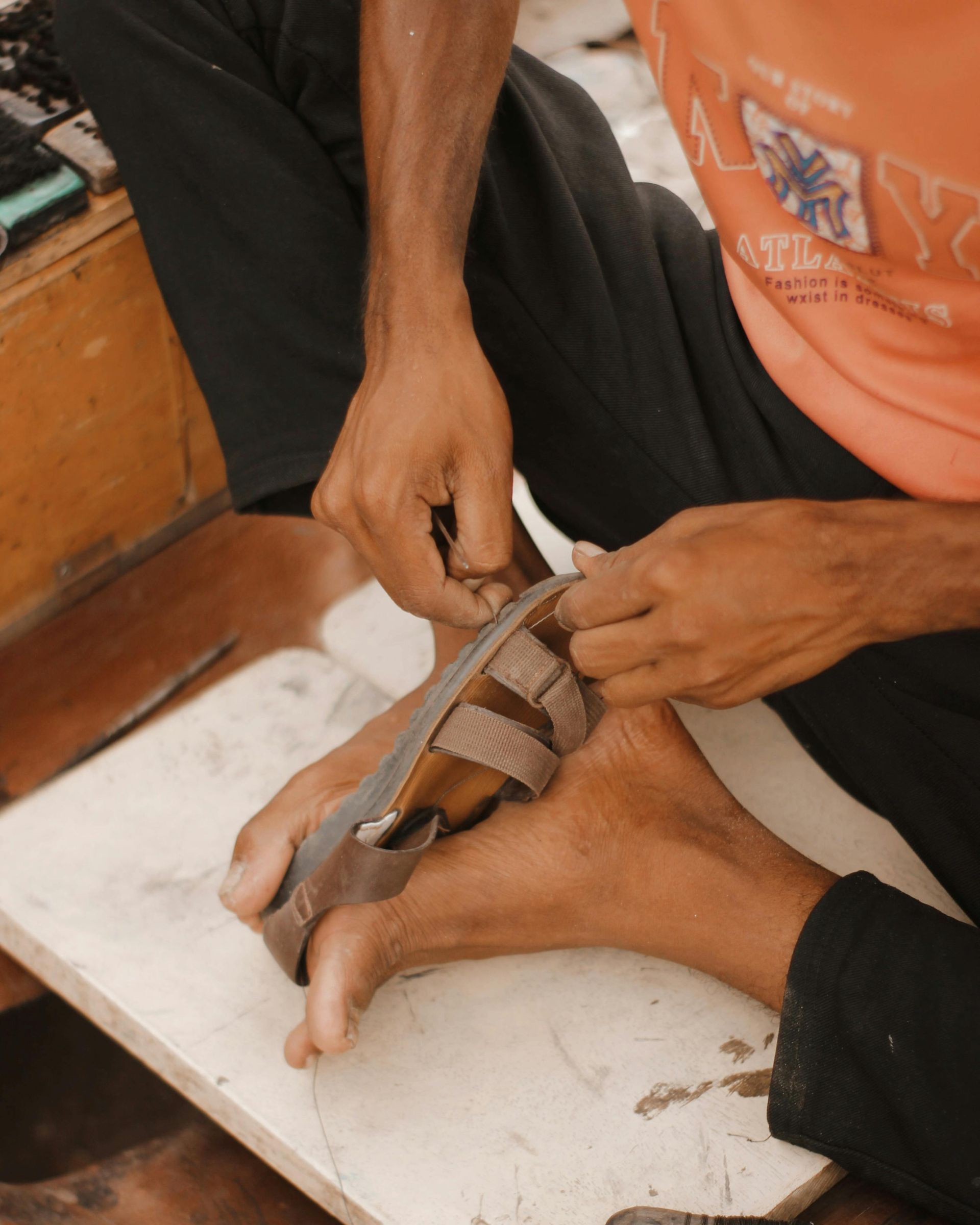 Person making a sandal, seated, using hands to work on leather straps over their foot, on a white surface.