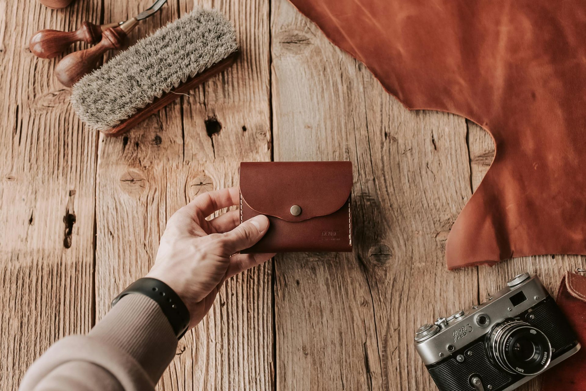 Hand holding a brown leather wallet on a wooden surface with tools and leather pieces.