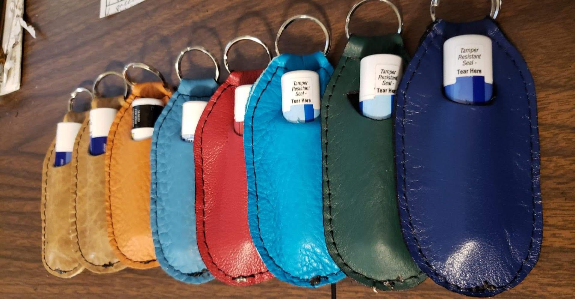 A row of eight colorful leather lip balm keychain holders in a gradient from tan to deep blue, resting on a wooden desk.