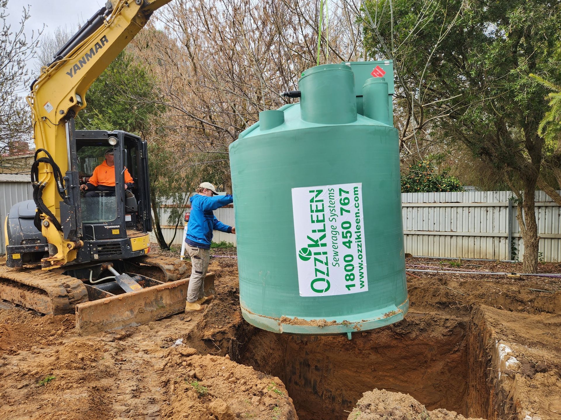 A backhoe and a person installing a large green septic tank in a muddy yard. — Mi Plumber Wagga Wagga in Lake Albert, NSW