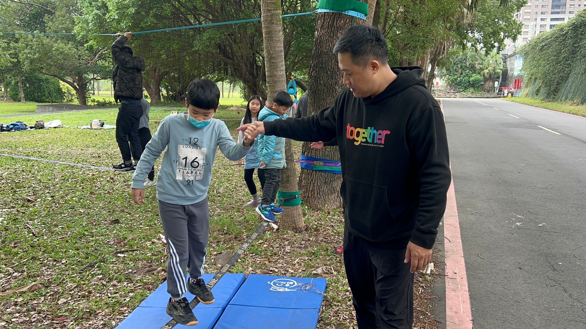 A man is helping a child balance on a blue mat in a park.