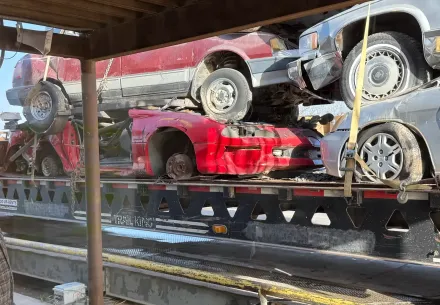A multi-level car carrier truck transporting several salvaged or dismantled vehicles, including red and gray cars.