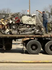 A person stands on the back of a flatbed truck, securing a large pile of crushed vehicles with a strap.