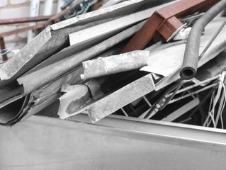 A pile of scrap metal, including gray sheet metal, a rusted red square beam, and a dark rubber hose in a metal container.