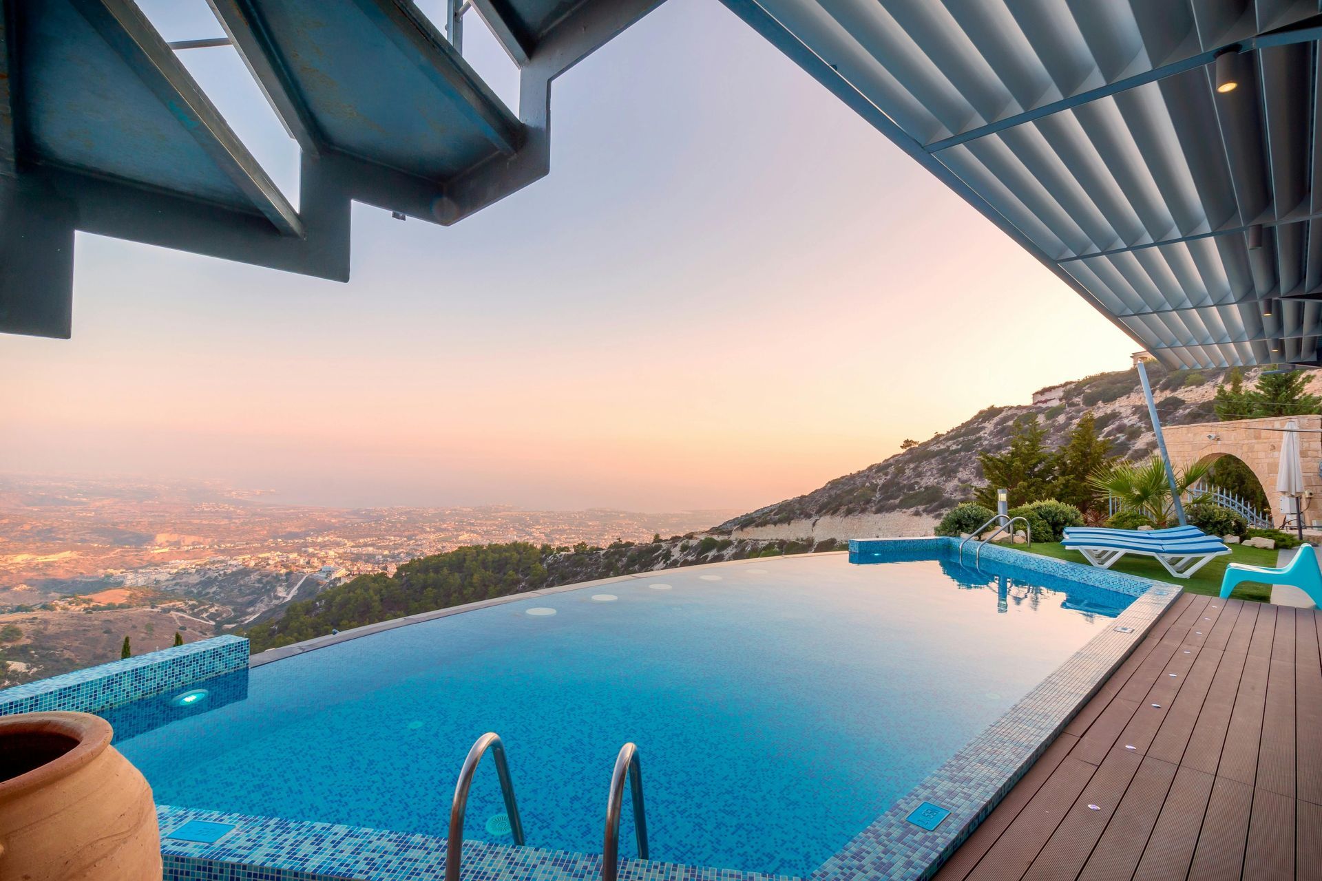 Infinity pool overlooks a city at sunset. Blue water, wood deck, and modern architecture.