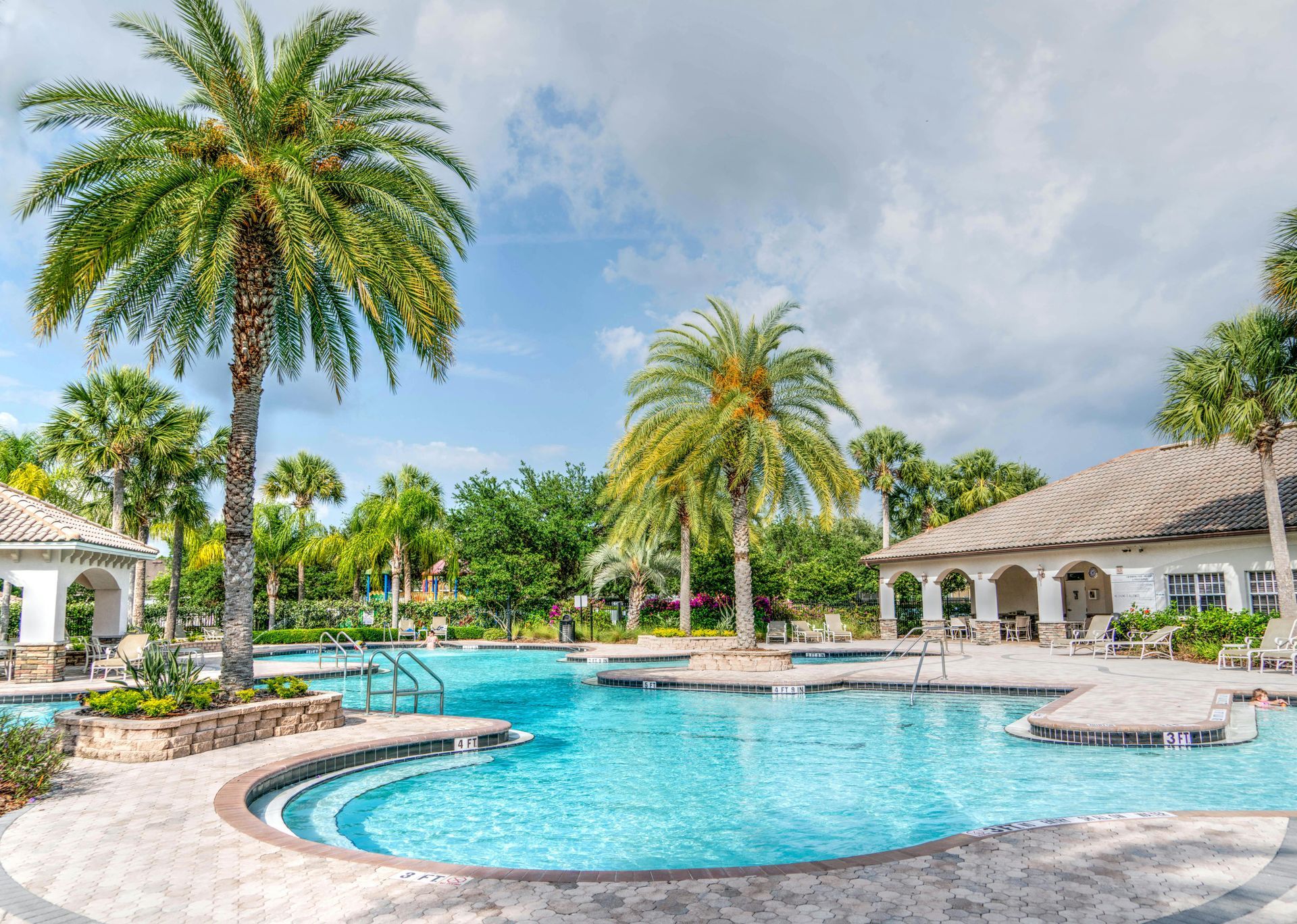 Pool area with palm trees, turquoise water, and a building with arches, under a blue sky.