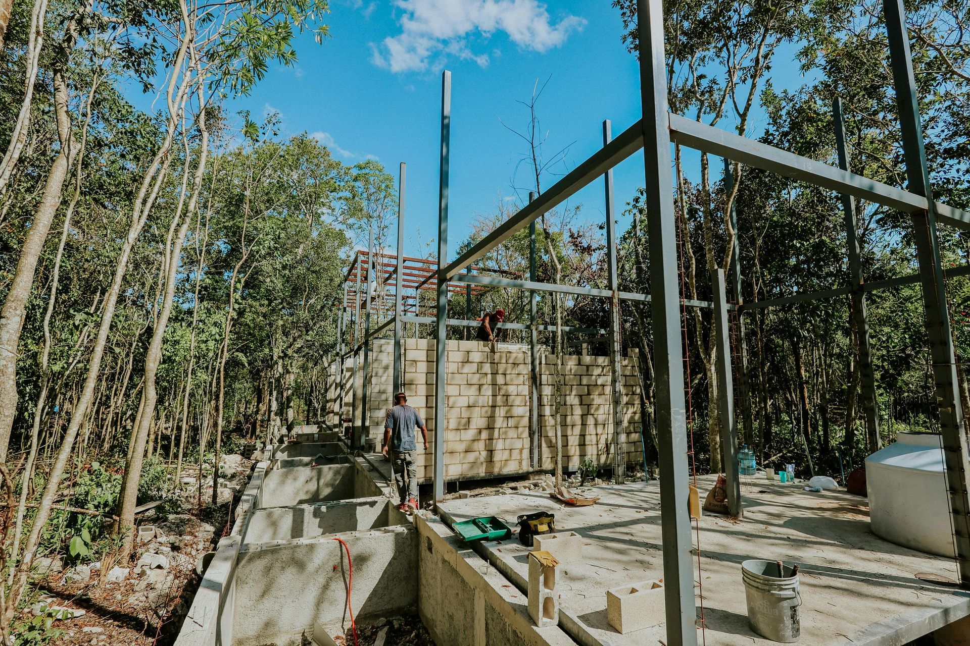 Construction site in a forest: metal frame of a building, man working, concrete structures, trees, blue sky.
