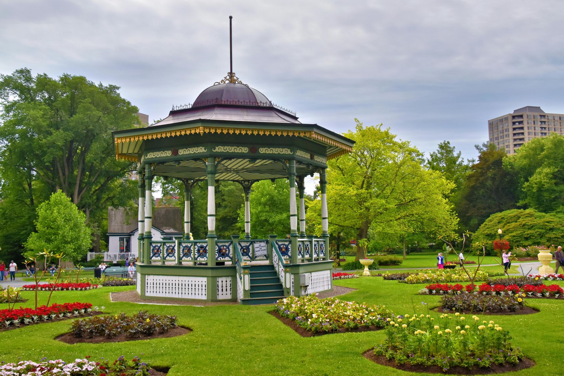 Green and gold bandstand in a park, surrounded by flowerbeds, trees, and a building. Cloudy sky.