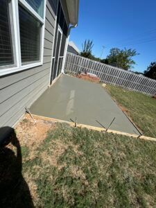 A new concrete patio is poured next to a house with a gray siding, on a sloped green lawn.