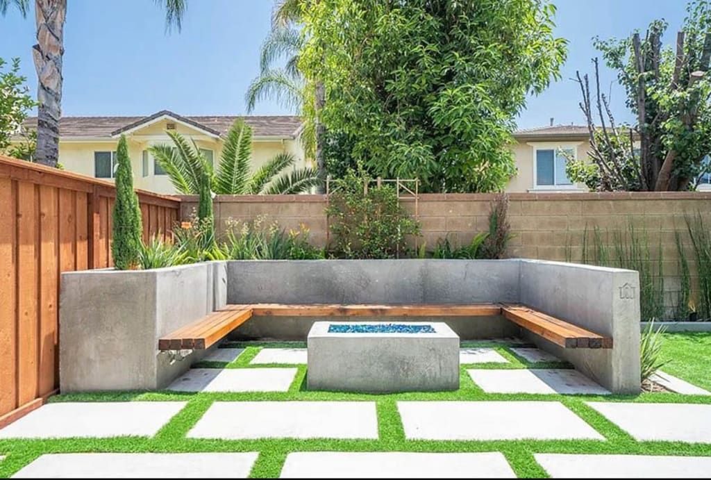 Concrete bench seating surrounding a fire pit on a patio with grass squares, adjacent to a fence.