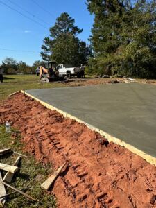 Freshly poured concrete slab with wooden edges, surrounded by reddish soil, a truck, and trees.