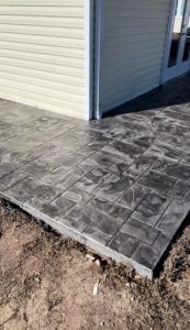 Stamped concrete patio, gray stone pattern, next to a beige house with tan siding.