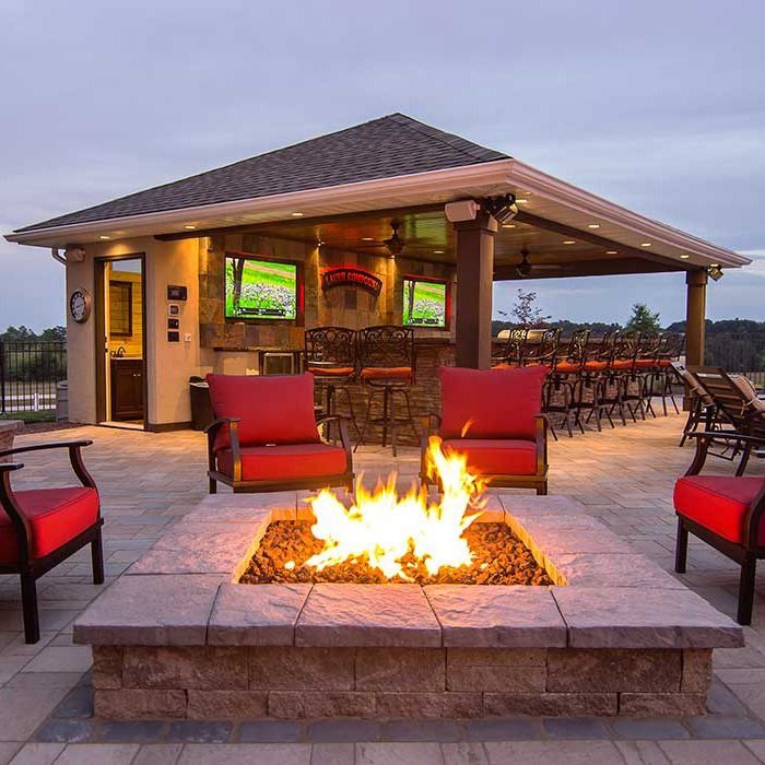 Outdoor patio with fire pit, bar, seating, and TVs. Red chairs around fire.