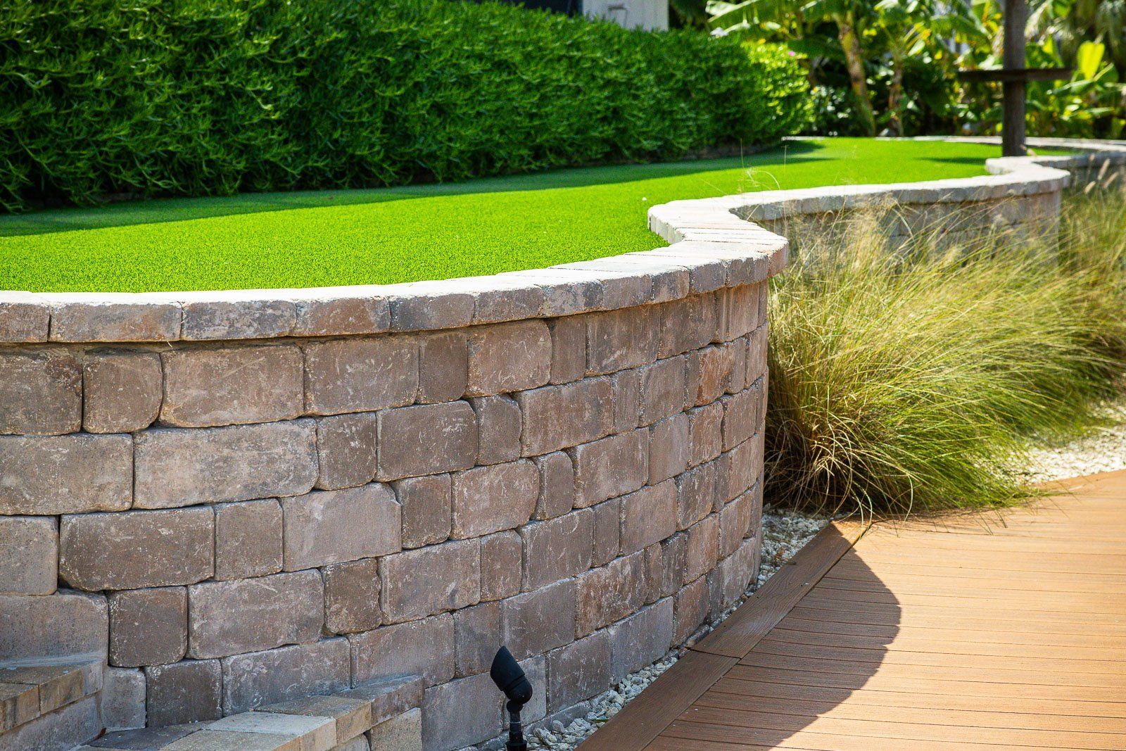 Curved retaining wall with brown bricks supports green grass and a shrub border.