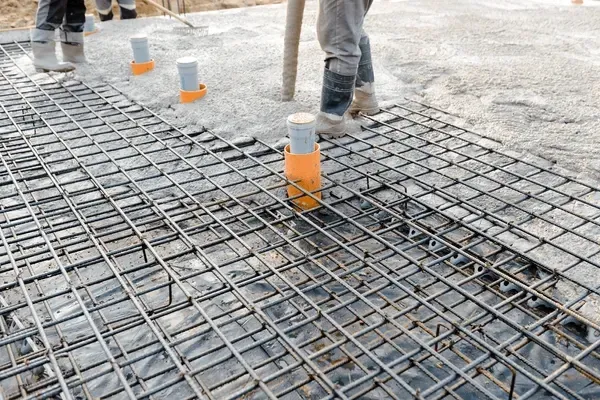 Construction site. Rebar grid laid on dark sheet, orange pipes, workers pouring concrete.